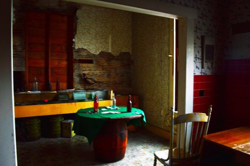 An abandoned kitchen with a green table, wooden chairs, and bottles, featuring peeling wallpaper and dim lighting.
