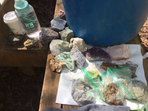 A wooden table with various colorful rocks, a water bottle, a coffee cup, and bags of smaller mineral samples.