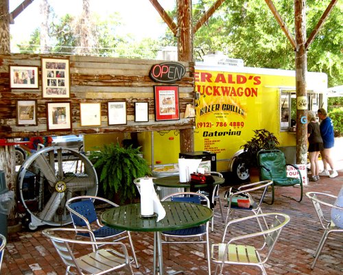 A food truck under a wooden shelter with tables and chairs, featuring a sign that says "Gerald's Truckwagon."