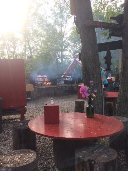 A rustic outdoor setting with a red table, flowers, and a barbecue area surrounded by trees and smoke.