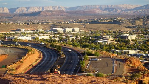 A winding road leads through a valley with mountains in the background, overlooking a small town and parking area.