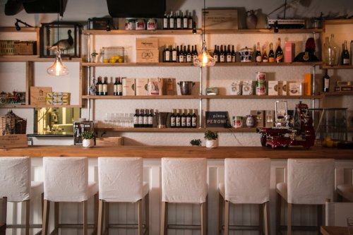 A cozy bar with wooden shelves displaying bottles and jars, and white bar stools lined up in front.