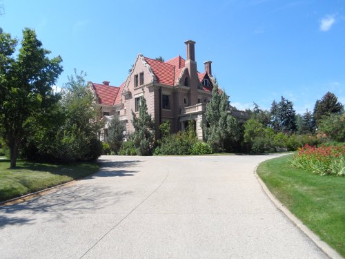 A large, elegant mansion with a red roof, surrounded by lush greenery and a winding driveway under a clear blue sky.