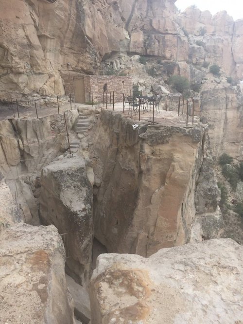 A rocky cliff with a narrow path and metal railings, leading to a small structure on the edge of the precipice.