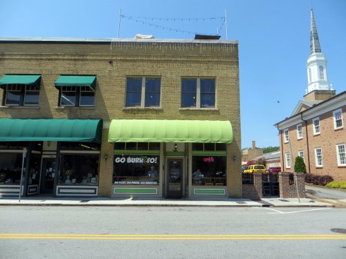 A street view of a brick building with green awnings, featuring a restaurant sign and a church steeple in the background.