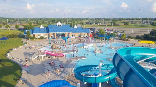 Aerial view of a water park with slides, a pool, and people enjoying the sunny day. Green lawns and blue skies in the background.