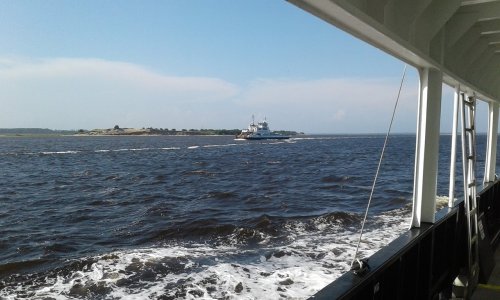 A view from a ferry showing a boat on dark waters with a distant shoreline under a clear sky.