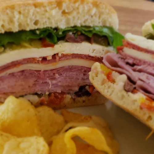 A close-up of a layered sandwich with meats, cheese, lettuce, and pickles, served with a side of potato chips.
