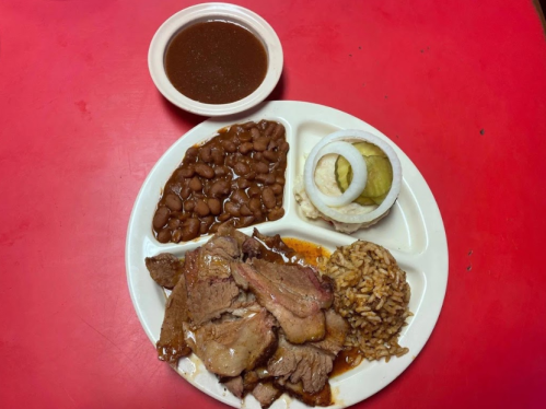 A plate of sliced meat with rice, beans, pickles, and onions, accompanied by a small bowl of sauce.