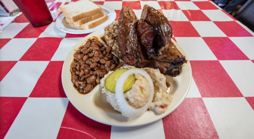 A plate of barbecue ribs, baked beans, pickles, and a side of bread on a red and white checkered tablecloth.