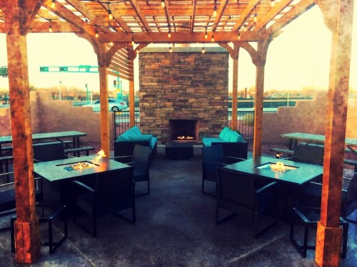 Cozy outdoor seating area with a stone fireplace, surrounded by tables and chairs under a wooden pergola.
