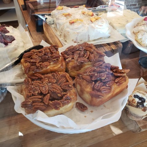 Four sticky buns topped with caramelized pecans, displayed in a bakery case with other pastries in the background.