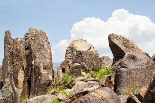 A cluster of large, weathered rocks with ancient petroglyphs, set against a blue sky with fluffy white clouds.