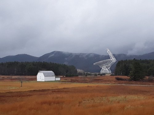 A white barn stands in a field with a large radio telescope and mountains in the background under a cloudy sky.