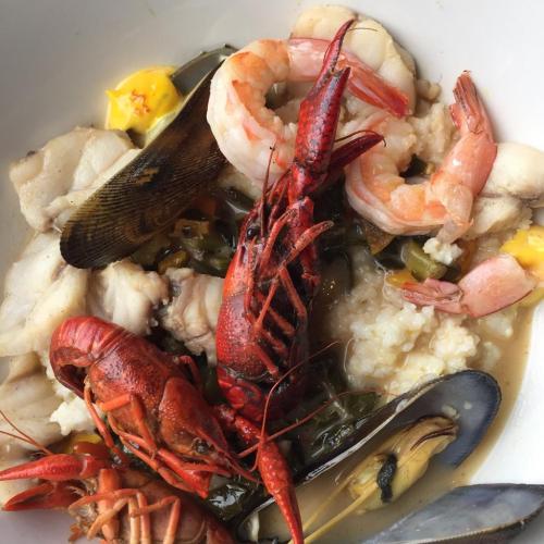 A bowl of seafood stew featuring shrimp, crawfish, fish, mussels, and greens in a creamy broth.