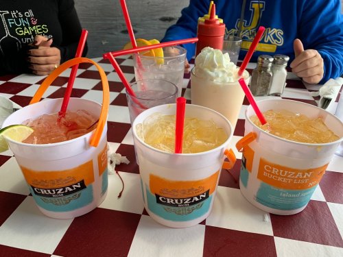 Four colorful drinks in large cups with straws on a checkered table, with condiments and glasses in the background.
