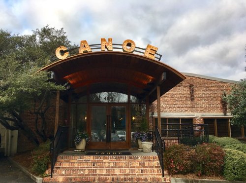 Entrance of a restaurant named "Canoe," featuring a wooden arch and brick facade, surrounded by greenery.