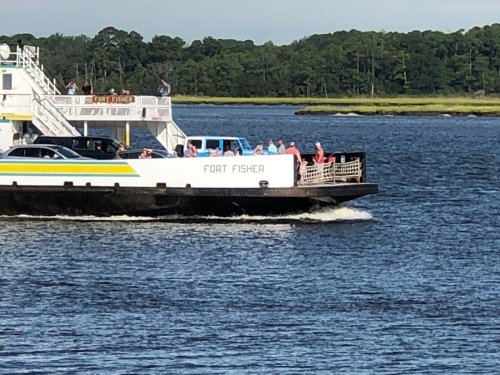 A ferry named "Fort Fisher" carries passengers across a calm waterway, surrounded by greenery.