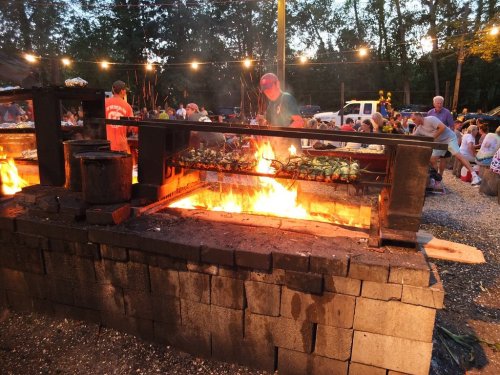 A lively outdoor gathering with people enjoying food near a large grill with flames and string lights in the background.