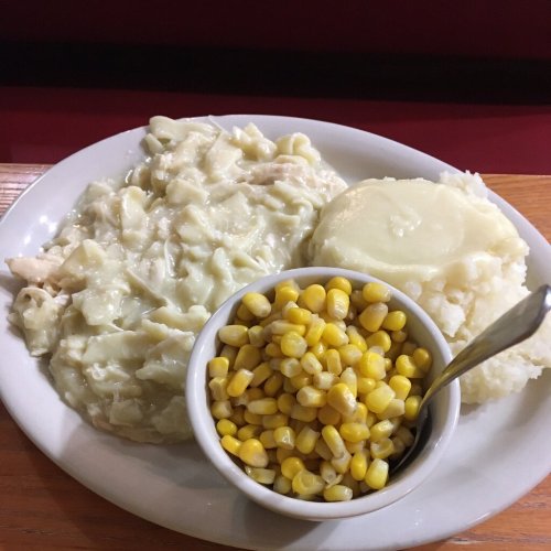 A plate of creamy chicken and noodles, mashed potatoes with gravy, and a side of corn in a bowl.