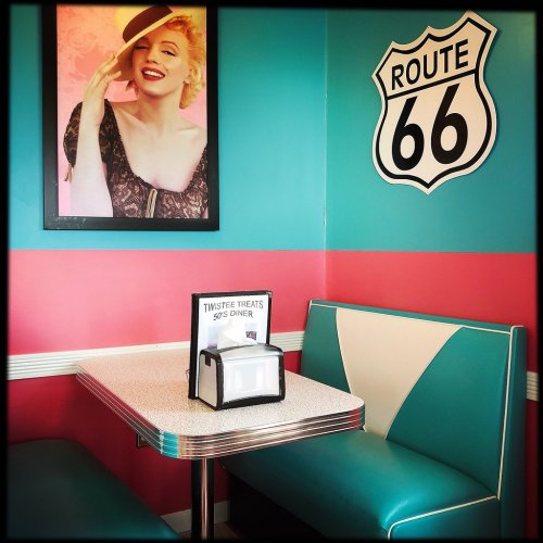 A retro diner booth with teal and pink walls, featuring a Route 66 sign and a vintage photo of a smiling woman.