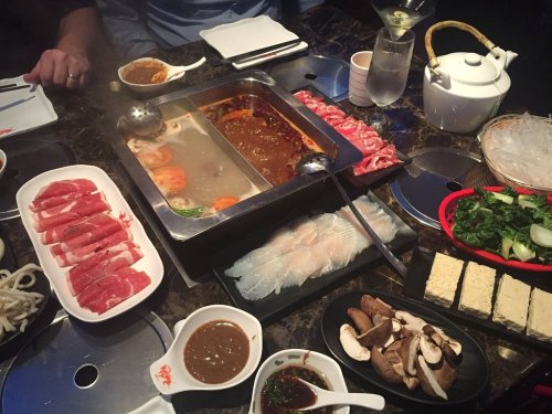 A table set for hot pot with various ingredients: sliced meats, vegetables, noodles, and dipping sauces.