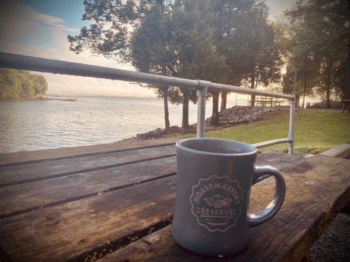 A coffee mug sits on a wooden table by a serene lakeside at sunrise, surrounded by trees and calm water.