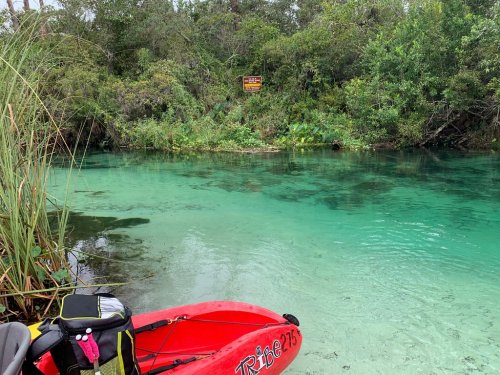 A red kayak is parked by a clear, turquoise river surrounded by lush greenery and a warning sign in the background.