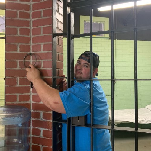 A man in a blue shirt holds a key while standing by a jail cell door in a prison setting.