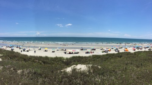 A panoramic view of a sunny beach with colorful umbrellas, people relaxing, and gentle waves in the background.
