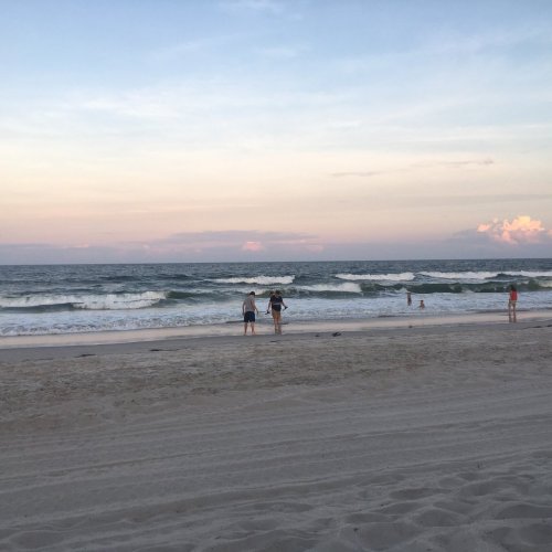 Two people walk along a sandy beach, with gentle waves and a pastel sky at sunset in the background.