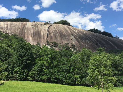 A large, smooth rock formation rises above lush green trees under a bright blue sky with scattered clouds.