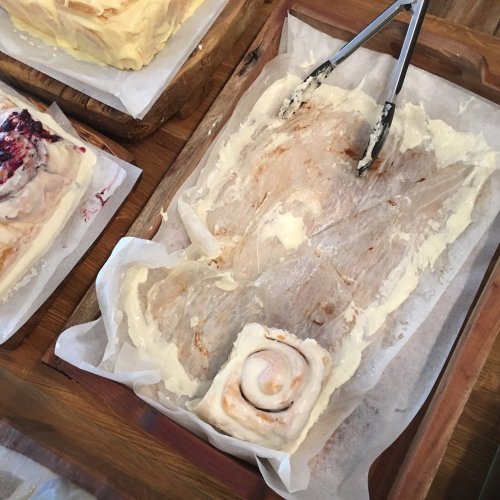 A partially eaten cinnamon roll on a wooden tray, surrounded by cream cheese frosting and other baked goods.