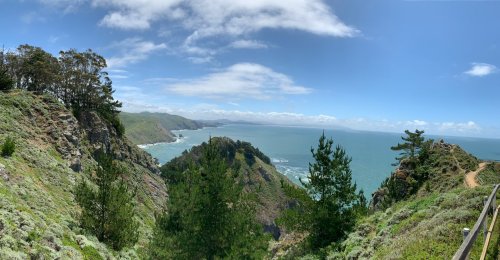 Panoramic view of a coastal landscape with cliffs, trees, and a blue ocean under a partly cloudy sky.