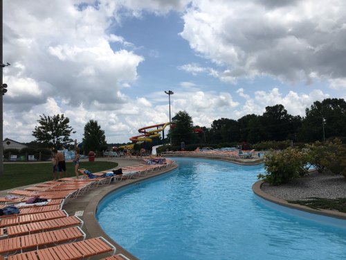 A winding lazy river pool with orange lounge chairs, surrounded by greenery and cloudy skies. Water slides in the background.