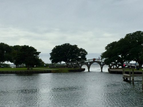 A scenic view of a bridge arching over a calm waterway, surrounded by trees under a cloudy sky.