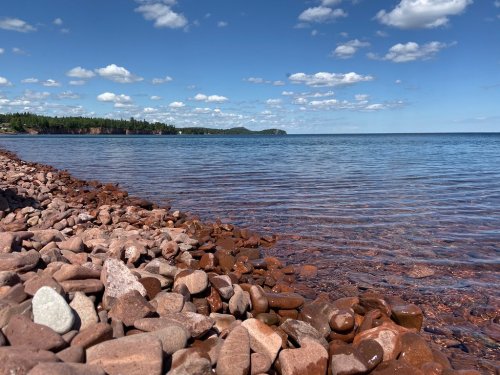 A rocky shoreline with smooth stones, clear blue water, and a bright sky with fluffy clouds in the background.