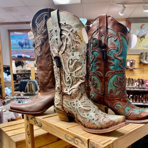 Three pairs of decorative cowboy boots displayed on a wooden shelf in a store, featuring various colors and designs.