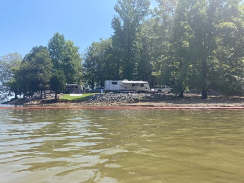 A view of a lakeside campsite with RVs and trees, seen from the water on a sunny day.