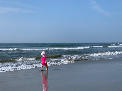 A child in a pink outfit walks along the shore, with gentle waves lapping at their feet under a clear blue sky.