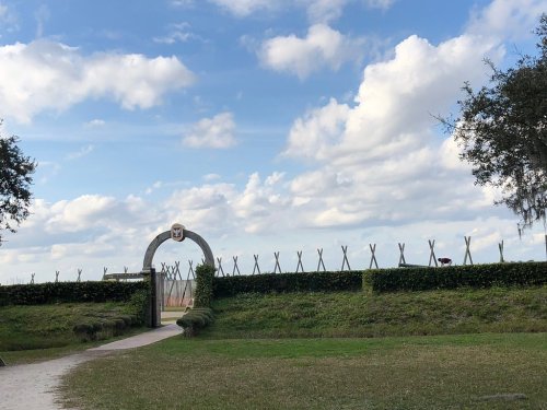 A stone archway leads to a grassy area with a fence and cloudy blue sky in the background.