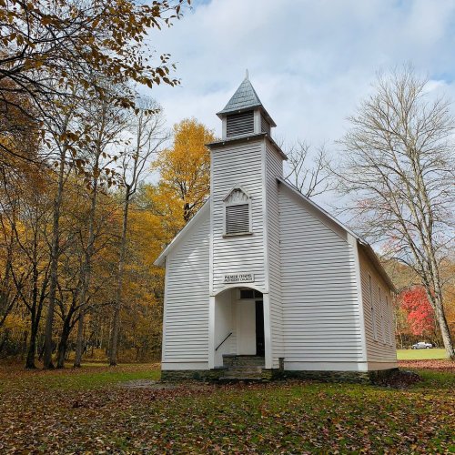 A small white church with a steeple, surrounded by autumn trees with colorful leaves under a cloudy sky.