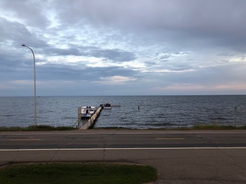 A calm lake scene with a wooden dock extending into the water under a cloudy sky, viewed from a roadside.