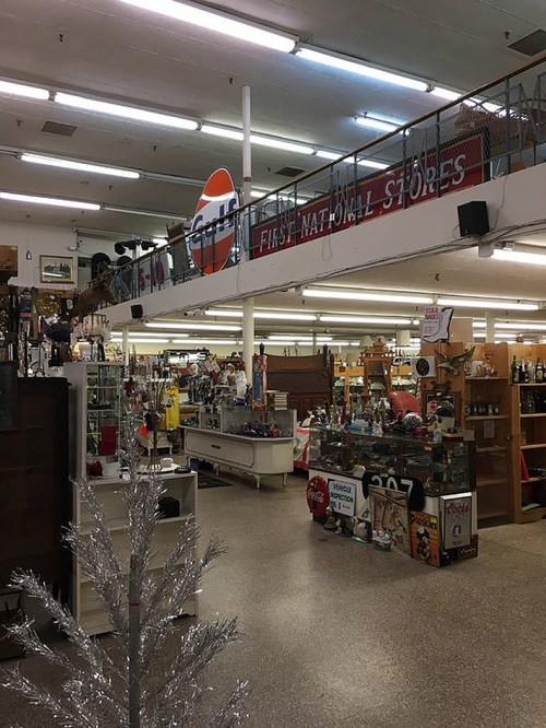 Interior of a vintage store with shelves of various items, a sign reading "First National Stores," and a silver tree.