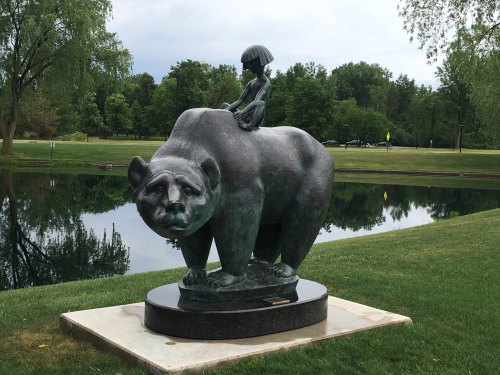 A bronze sculpture of a bear with a child holding an umbrella sitting on its back, set in a park by a pond.