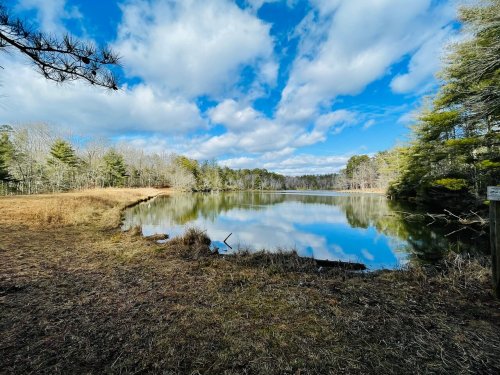 A serene lake surrounded by trees under a partly cloudy sky, reflecting the landscape on its calm surface.