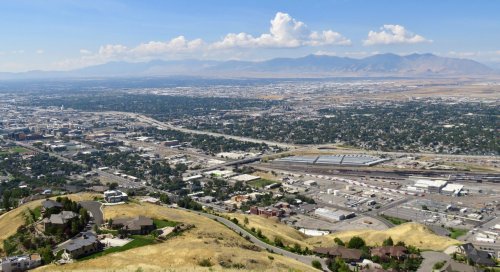 Aerial view of a city with mountains in the background, featuring green hills and urban development.
