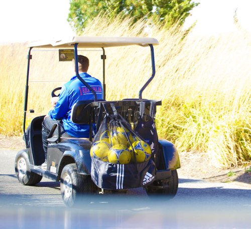 A person in a blue jacket drives a golf cart loaded with soccer balls in a grassy area.