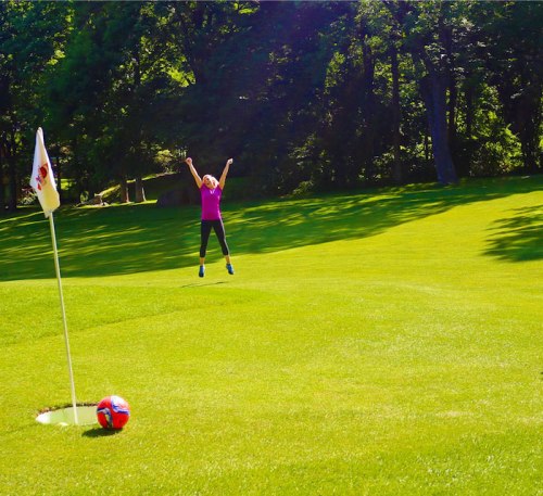A person in a pink shirt jumps joyfully on a green field near a soccer ball and a flag.