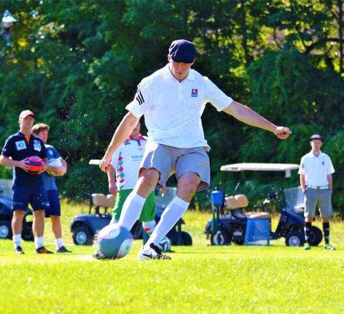 A man in a white shirt and cap kicks a soccer ball on a grassy field, with spectators and golf carts in the background.
